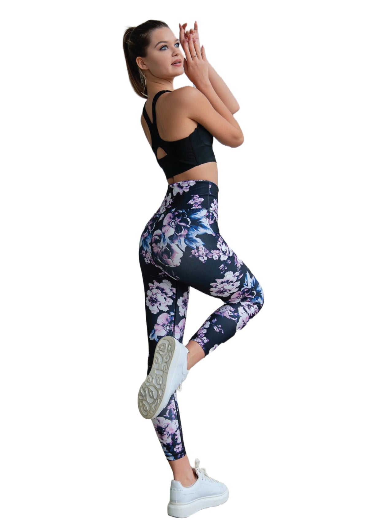 Person in einem schwarzen Sport-BH, die in einer Yoga-Pose auf einem Bein steht und Lismina-Leggings mit Blumenmuster und weiße Turnschuhe trägt.